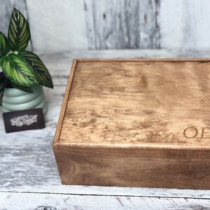 May include: A wooden box with a lid, stained in a warm brown color. The initials "OEB" are engraved on the front. A small potted plant with green and pink striped leaves sits to the left of the box.