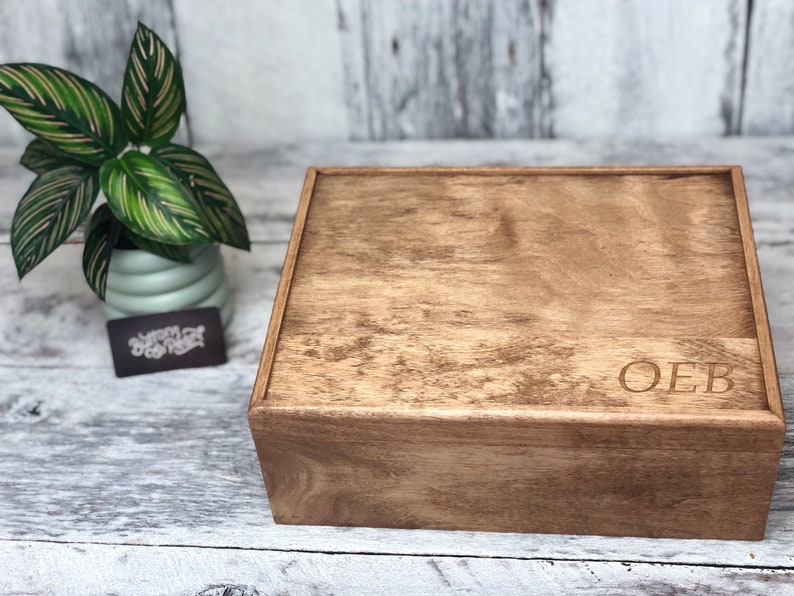 May include: A wooden box with a lid, stained in a warm brown color. The initials "OEB" are engraved on the front. A small potted plant with green and pink striped leaves sits to the left of the box.