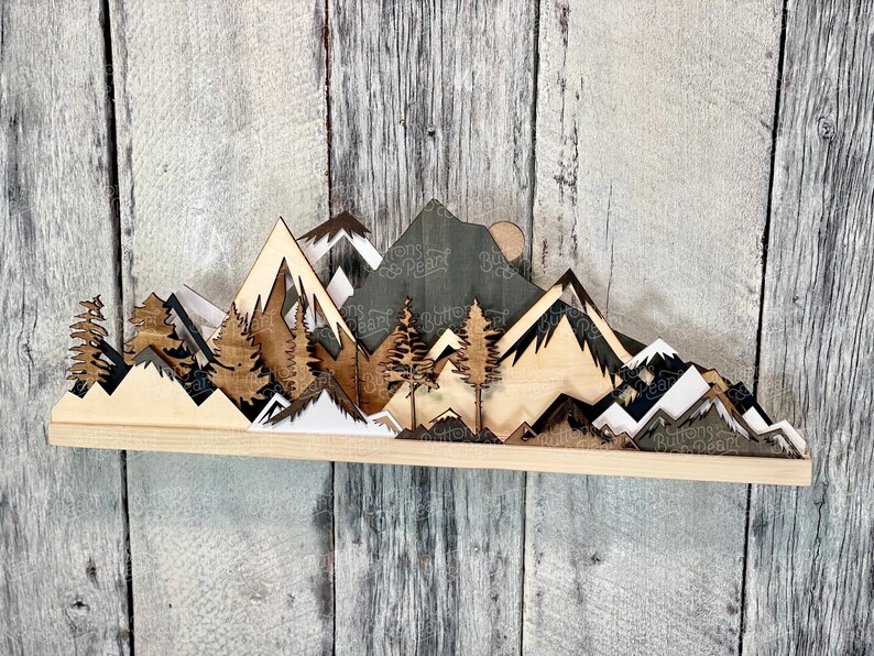 May include: Wooden wall art featuring a layered mountain range scene with trees and a sun. The mountains are in shades of brown, white, and black. The trees are brown and the sun is a light brown circle.
