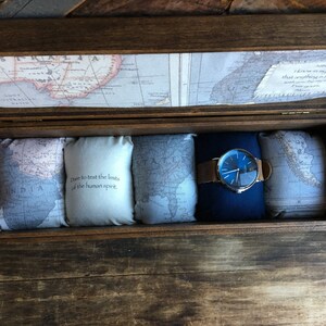 Puede incluir: Una caja de madera con un dise&ntilde;o de mapa en la tapa. La caja est&aacute; abierta y contiene cuatro peque&ntilde;as almohadas con mapas en ellas y un reloj con una esfera azul y una correa de cuero marr&oacute;n. Las almohadas tienen el texto "Dare to test the limits of the human spirit" impreso en ellas.