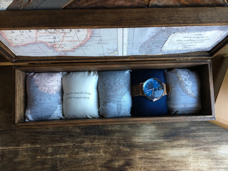 Puede incluir: Una caja de madera con un dise&ntilde;o de mapa en la tapa. La caja est&aacute; abierta y contiene cuatro peque&ntilde;as almohadas con mapas en ellas y un reloj con una esfera azul y una correa de cuero marr&oacute;n. Las almohadas tienen el texto "Dare to test the limits of the human spirit" impreso en ellas.