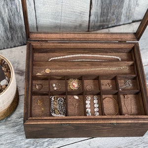 May include: A dark brown wooden jewelry box with a glass lid. The box has multiple compartments for storing jewelry. The box is open and the compartments are filled with various pieces of jewelry, including necklaces, earrings, and bracelets.