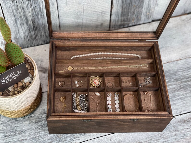 May include: A dark brown wooden jewelry box with a glass lid. The box has multiple compartments for storing jewelry. The box is open and the compartments are filled with various pieces of jewelry, including necklaces, earrings, and bracelets.
