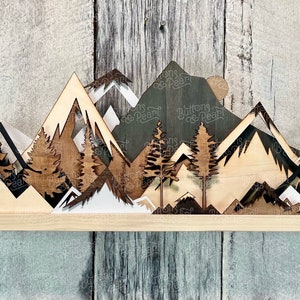 May include: A wooden mountain range wall decor with a layered design. The mountains are painted in shades of brown, black, and white. The piece is mounted on a wooden shelf.