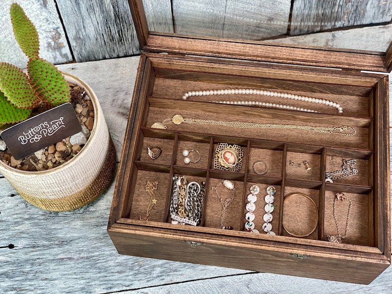 May include: A wooden jewelry box with a glass lid, divided into compartments, filled with various jewelry pieces, including necklaces, earrings, bracelets, and rings. The box is labeled "Buttons & Pearl".