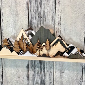 May include: A wooden wall art piece depicting a mountain range with trees and a moon. The mountains are layered and have different shades of brown, black, and white. The trees are dark brown and the moon is a light brown.