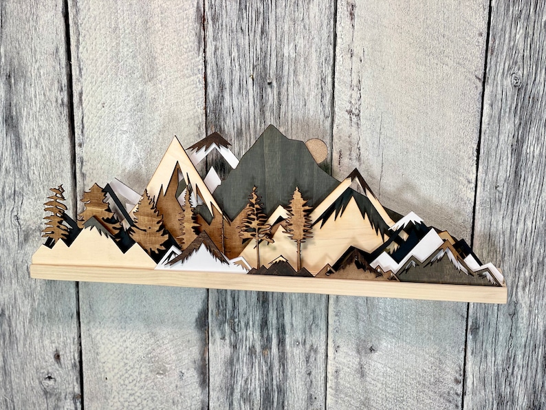 May include: A wooden wall art piece depicting a mountain range with trees and a moon. The mountains are layered and have different shades of brown, black, and white. The trees are dark brown and the moon is a light brown.