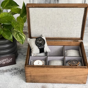 May include: A wooden watch box with a grey fabric lining and six compartments for storing watches. The box is open and there are three watches inside, two with black faces and one with a gold face.