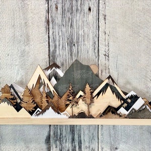 May include: A wooden wall art piece depicting a mountain range with a sunset in the background. The mountains are layered and have different shades of brown, white, and black. There are also small trees in the foreground.