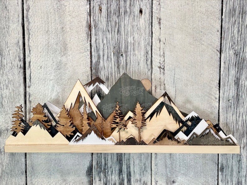 May include: A wooden wall art piece depicting a mountain range with a sunset in the background. The mountains are layered and have different shades of brown, white, and black. There are also small trees in the foreground.