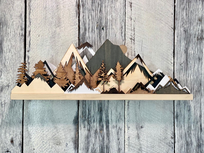 May include: Wooden wall art depicting a mountain range with trees and a sun. The mountains are layered and cut out in different shades of brown, black, and white. The trees are dark brown and the sun is a light brown circle.