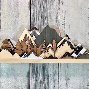 May include: A wooden wall art piece depicting a mountain range with trees and a moon. The mountains are layered and cut out in different shades of brown, black, and white.