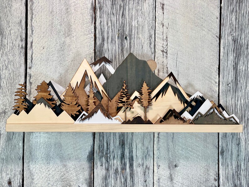 May include: A wooden wall art piece depicting a mountain range with trees and a moon. The mountains are layered and cut out in different shades of brown, black, and white.