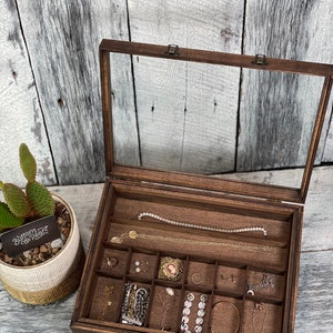 May include: A wooden jewelry box with a glass top and multiple compartments. The box is filled with various jewelry pieces, including necklaces, earrings, and bracelets. The box has a label that reads "Buttons & Pearl".