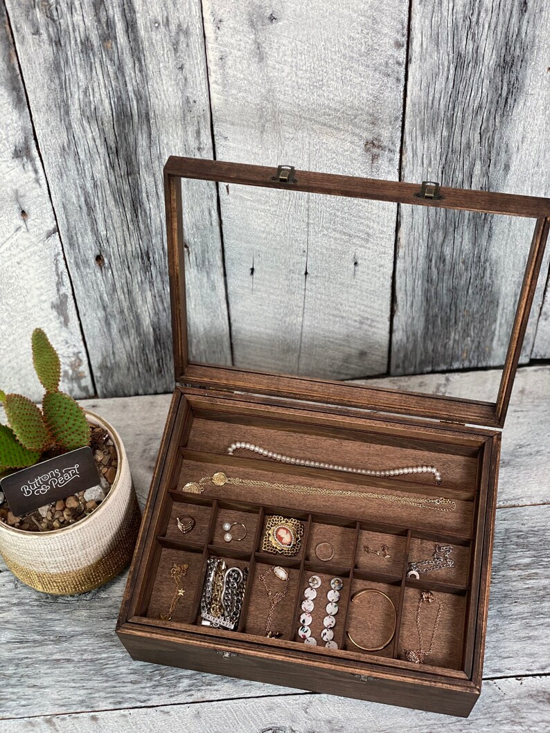 May include: A wooden jewelry box with a glass top and multiple compartments. The box is filled with various jewelry pieces, including necklaces, earrings, and bracelets. The box has a label that reads "Buttons & Pearl".