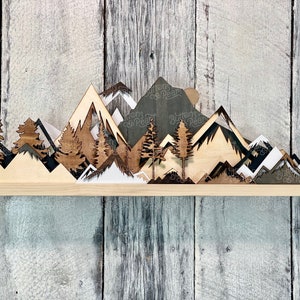 May include: Wooden wall decor featuring a layered mountain range silhouette with trees and snow-capped peaks. The mountains are painted in shades of brown, black, and white.