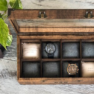 Puede incluir: Una caja de madera para relojes con tapa de cristal y ocho compartimentos. La caja tiene un tinte marr&oacute;n y bisagras de lat&oacute;n. Los compartimentos est&aacute;n forrados con tela gris. Dos relojes son visibles en la caja. Un reloj tiene una correa negra y una esfera blanca. El otro reloj tiene una correa marr&oacute;n y una esfera dorada. La caja tiene una peque&ntilde;a almohada blanca en cada compartimento con texto que dice "When you are more with each second that goes by" y "40&deg;47'14"N 73&deg;40'22"W".