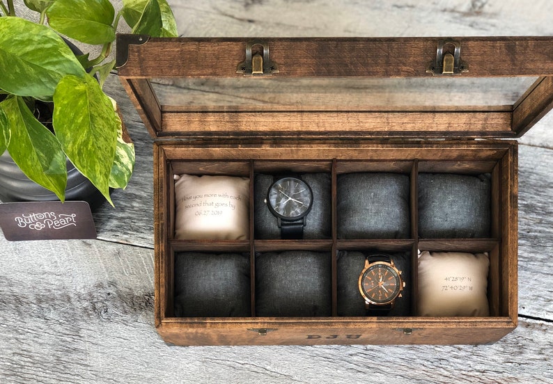 Puede incluir: Una caja de madera para relojes con tapa de cristal y ocho compartimentos. La caja tiene un tinte marr&oacute;n y bisagras de lat&oacute;n. Los compartimentos est&aacute;n forrados con tela gris. Dos relojes son visibles en la caja. Un reloj tiene una correa negra y una esfera blanca. El otro reloj tiene una correa marr&oacute;n y una esfera dorada. La caja tiene una peque&ntilde;a almohada blanca en cada compartimento con texto que dice "When you are more with each second that goes by" y "40&deg;47'14"N 73&deg;40'22"W".