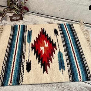 May include: A white woven rug with a geometric pattern in shades of red, blue, and black. The rug has fringe on all sides and is perfect for adding a touch of Southwestern style to your home.