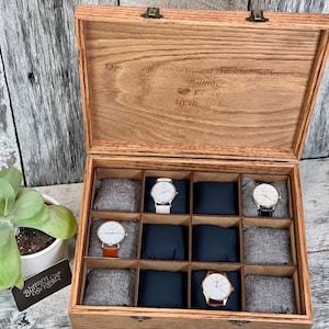 May include: Wooden watch box with a hinged lid, featuring compartments for multiple watches. The box is open, revealing several watches with different colored bands. The lid has engraved text. A succulent plant in a white pot is on the left.