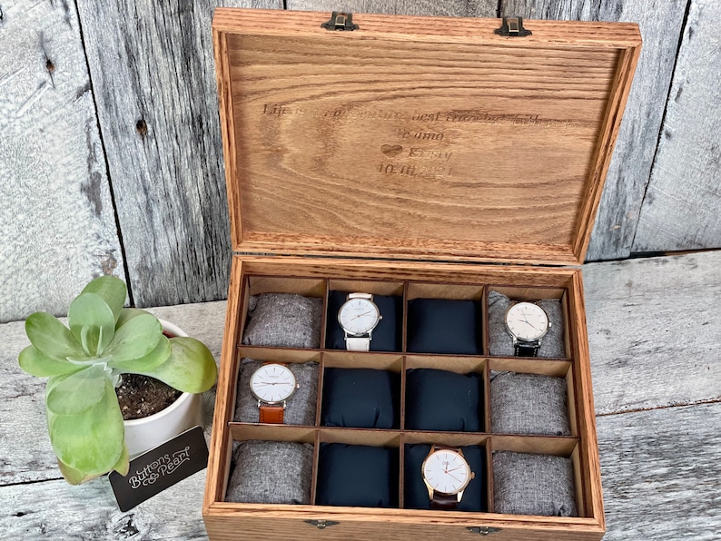 May include: Wooden watch box with a hinged lid, featuring compartments for multiple watches. The box is open, revealing several watches with different colored bands. The lid has engraved text. A succulent plant in a white pot is on the left.
