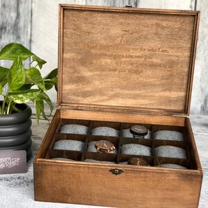 May include: Wooden watch box with a hinged lid, featuring a dark brown finish and a compartmented interior with gray cushions. The lid has an engraved message. A potted plant and a business card are in the background.