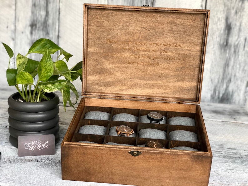 May include: Wooden watch box with a hinged lid, featuring a dark brown finish and a compartmented interior with gray cushions. The lid has an engraved message. A potted plant and a business card are in the background.