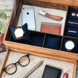 May include: A wooden box with two drawers, the top drawer has compartments for watches, a wallet, a knife, cufflinks, and a tie clip. The bottom drawer has a pen, glasses, a wallet, and a multi-tool. The box is labeled "Buttons & Pearl".