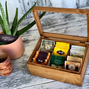 Puede incluir: Una caja de madera para té con tapa de vidrio, que contiene varias bolsitas de té. La caja está dividida en compartimentos y tiene una etiqueta que dice "Buttons & Pearl".