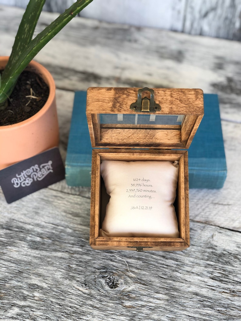 Puede incluir: Una caja de madera con tapa con bisagras y una peque&ntilde;a almohada blanca en su interior. La almohada tiene el texto "1624 d&iacute;as, 58.996 horas, 2.339.760 minutos. Y contando... J&A|12.21.19" impreso en ella.