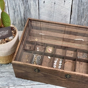 May include: A wooden jewelry box with a glass lid and a brown felt interior. The box has multiple compartments for storing rings, earrings, necklaces, and bracelets. The box is labeled "Buttons & Pearl".