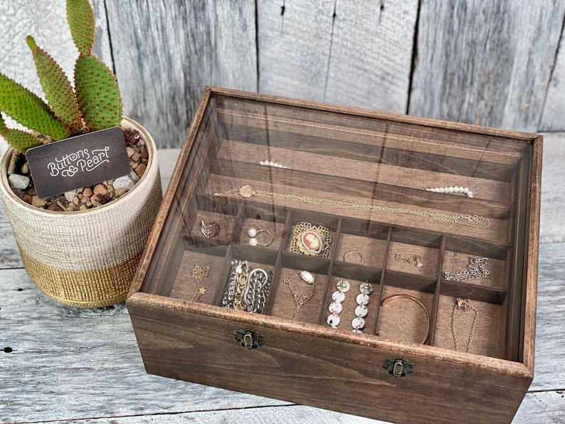 May include: A wooden jewelry box with a glass lid and a brown felt interior. The box has multiple compartments for storing rings, earrings, necklaces, and bracelets. The box is labeled "Buttons & Pearl".