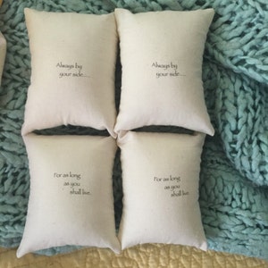 Puede incluir: Cuatro peque&ntilde;os cojines de tela blanca con texto bordado. Los cojines tienen el texto "Always by your side..." y "For as long as you shall live." Los cojines est&aacute;n dispuestos en dos filas de dos sobre una superficie azul claro y amarilla.