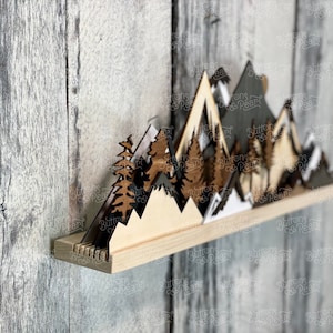 May include: Wooden mountain range wall decor with a shelf. The mountains are layered and painted in shades of brown, black, and white. The shelf is made of natural wood.