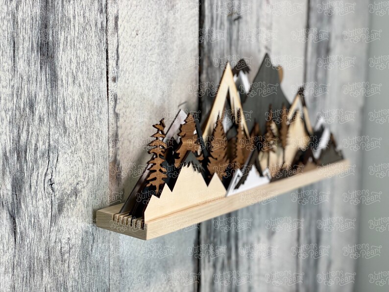 May include: Wooden mountain range wall decor with a shelf. The mountains are layered and painted in shades of brown, black, and white. The shelf is made of natural wood.