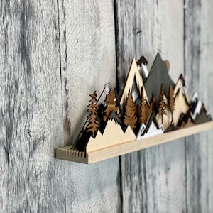 May include: A wooden wall shelf with a layered mountain range design. The mountains are painted in shades of brown, white, and gray. The shelf is mounted on a gray wood wall.