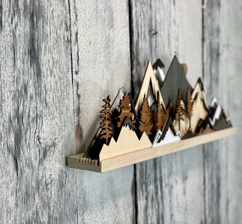 May include: A wooden wall shelf with a layered mountain range design. The mountains are painted in shades of brown, white, and gray. The shelf is mounted on a gray wood wall.