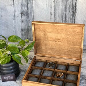 May include: Wooden watch box with a hinged lid, featuring 12 gray padded compartments. The box is open, revealing two watches. The lid is engraved with the text "3/10/20 My Hero". A potted plant is visible on the left.
