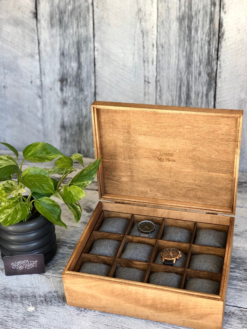 May include: Wooden watch box with a hinged lid, featuring 12 gray padded compartments. The box is open, revealing two watches. The lid is engraved with the text "3/10/20 My Hero". A potted plant is visible on the left.