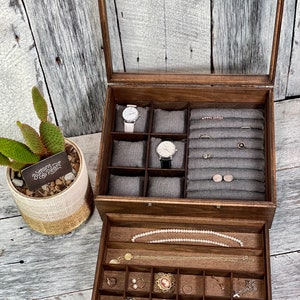 May include: A wooden jewelry box with a glass top and a pull-out drawer. The top compartment has multiple slots for watches and rings. The drawer has a variety of compartments for storing jewelry, including necklaces, earrings, and bracelets. The box is labeled "Buttons & Pearl".