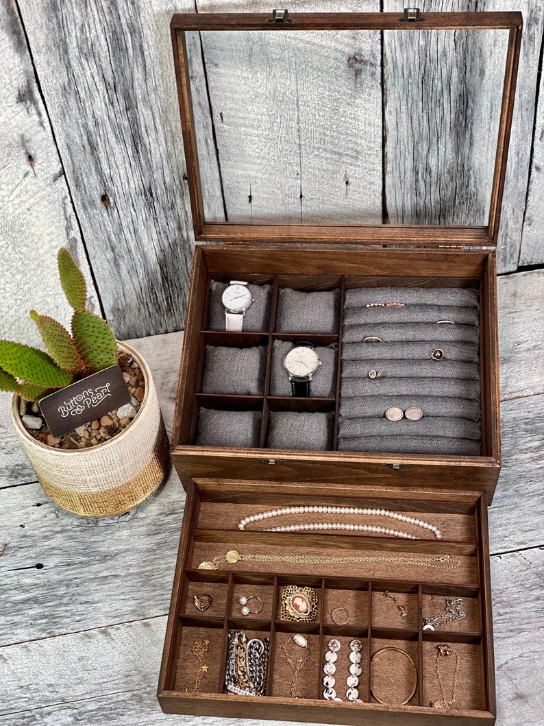 May include: A wooden jewelry box with a glass top and a pull-out drawer. The top compartment has multiple slots for watches and rings. The drawer has a variety of compartments for storing jewelry, including necklaces, earrings, and bracelets. The box is labeled "Buttons & Pearl".