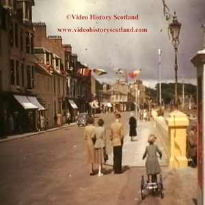 May include: A black and white photo of a street scene in a town. There are buildings on both sides of the street, and people are walking along the sidewalk. There are flags hanging from the buildings, and a street lamp is visible in the background. The text "Video History Scotland" and "www.videohistoryscotland.com" is visible in the image.