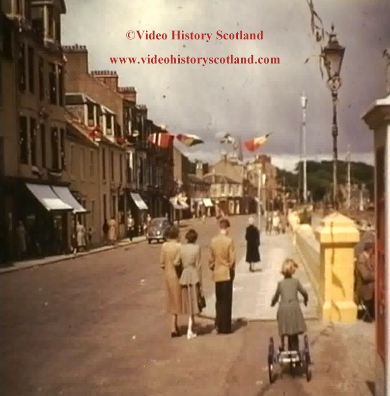 May include: A black and white photo of a street scene in a town. There are buildings on both sides of the street, and people are walking along the sidewalk. There are flags hanging from the buildings, and a street lamp is visible in the background. The text "Video History Scotland" and "www.videohistoryscotland.com" is visible in the image.