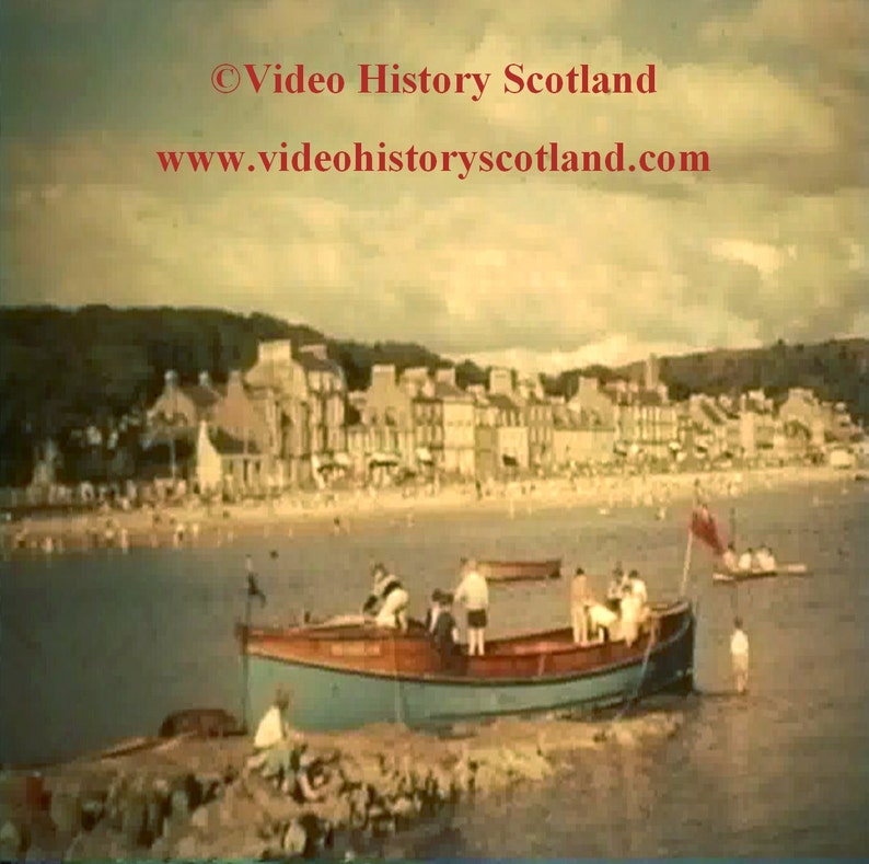 May include: A vintage photograph of a small boat docked at a beach with a town in the background. The boat is blue and red with people on board. The beach is crowded with people and the town is made up of several buildings.