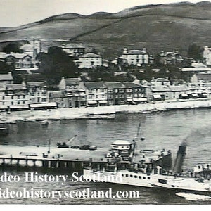 May include: A black and white photo of a town with a pier and a boat docked at the pier. The boat is a paddle steamer with a large smokestack. The town is built on a hillside and there are buildings in the background.