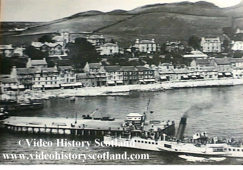 May include: A black and white photo of a town with a pier and a boat docked at the pier. The boat is a paddle steamer with a large smokestack. The town is built on a hillside and there are buildings in the background.