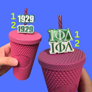 May include: Two pink cups with lids and straws. One cup has a green and white 1929 charm on the straw. The other cup has a green and white charm with the letters IΦA on the straw.