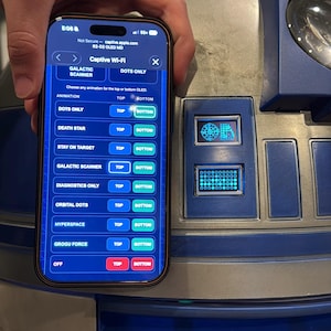 May include: A smartphone displaying a Captive Wi-Fi configuration menu for an R2-D2 unit, featuring animation options like Galactic Scanner and Death Star, held in front of a silver and blue droid dome with illuminated rectangular display panels.