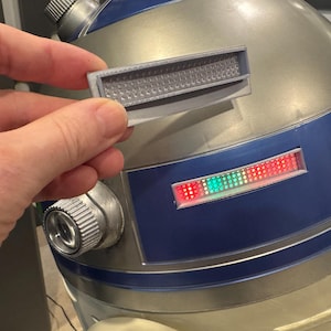 May include: A silver and blue robotic dome featuring a rectangular light panel with red and green LED dots. A separate grey rectangular component with a textured grid pattern is held in front of the dome, matching the design of the robot's features.