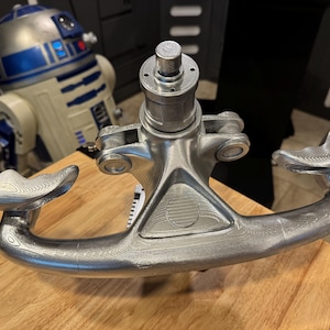 Op de afbeelding: Een zilverkleurige, 3D-geprinte stuurknuppel met een centrale cilindrische kolom met een knop aan de bovenkant en twee gebogen, ergonomische handgrepen aan weerszijden. Het oppervlak vertoont zichtbare horizontale lijnen van het printproces.